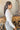 Side view of model wearing the On The Daily Bodysuit, that has white textured fabric, a high neck with lettuce trim, long sleeves with lettuce trim, and a thong bottom with hook closures. Worn with jeans.