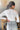 Back view of model wearing the On The Daily Bodysuit, that has white textured fabric, a high neck with lettuce trim, long sleeves with lettuce trim, and a thong bottom with hook closures. Worn with jeans.