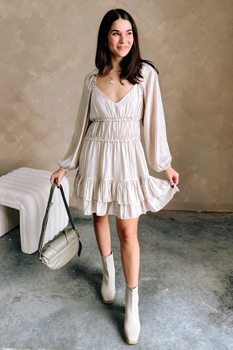 Full body view of model wearing the Taryn Beige Ruffle Tiered Mini Dress which features light taupe light weight fabric, light taupe lining, a two-tiered flare skirt, ruffle details, a mini length hem, a v-neckline, long sleeves with elastic wrists, and a