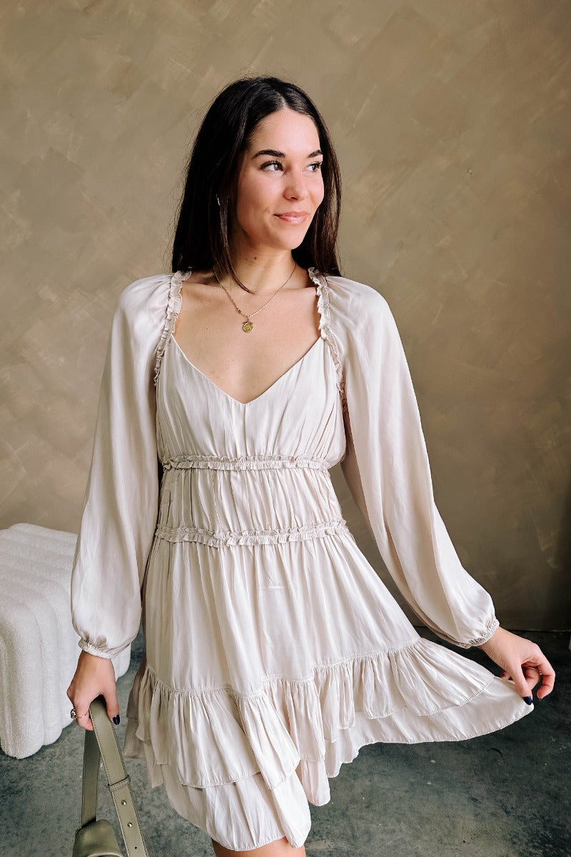 Front view of model wearing the Taryn Beige Ruffle Tiered Mini Dress which features light taupe light weight fabric, light taupe lining, a two-tiered flare skirt, ruffle details, a mini length hem, a v-neckline, long sleeves with elastic wrists, and a bac