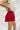 Side view of the Maeve Skort that features a red satin material, a thick elastic waist band with a tie detail, a ruffle tiered design, a shorts lining, and a mini length.