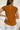 Back view of the Hampton Social Top that features a caramel material, a high rounded neckline with a ruffle hem, a short flowy sleeve, a pleated design, a lettuce edge bottom hem, and a flowy fit.
