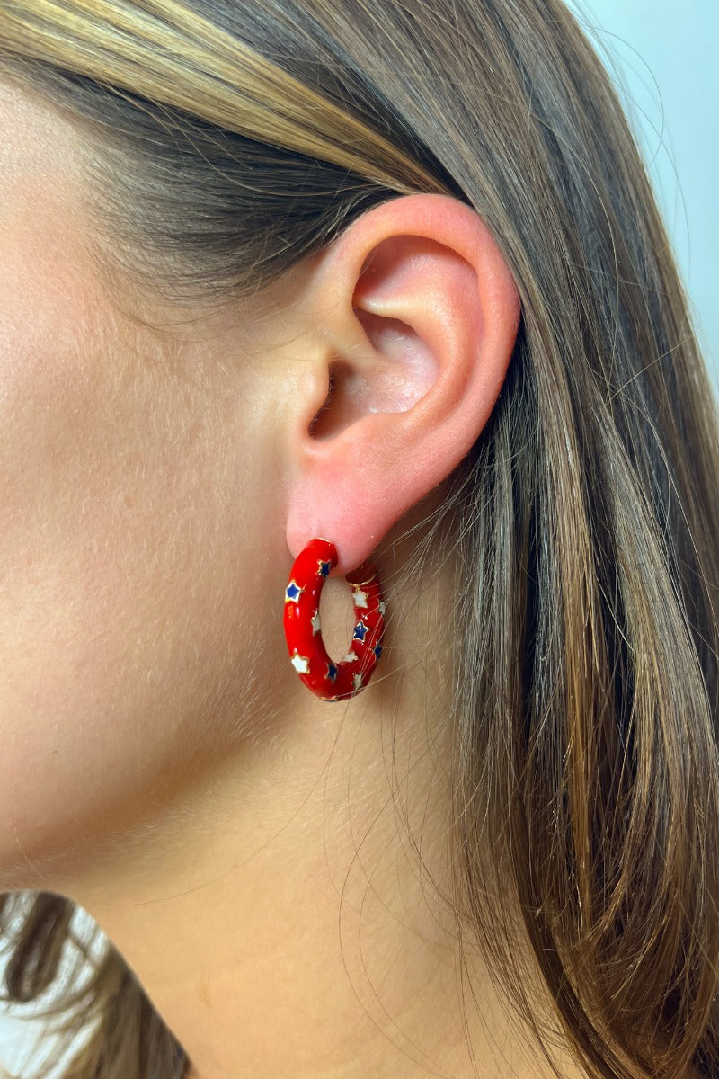 Side view of model wearing the Star Gazing Earrings in Red which features small open red hoop with white and blue stars design outlined in gold details.