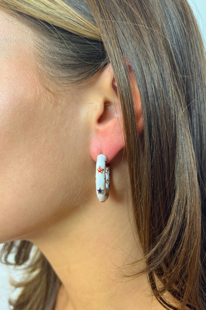 Side view of model wearing the Star Gazing Earrings in White which features small open white hoop with red and blue stars design outlined in gold details.