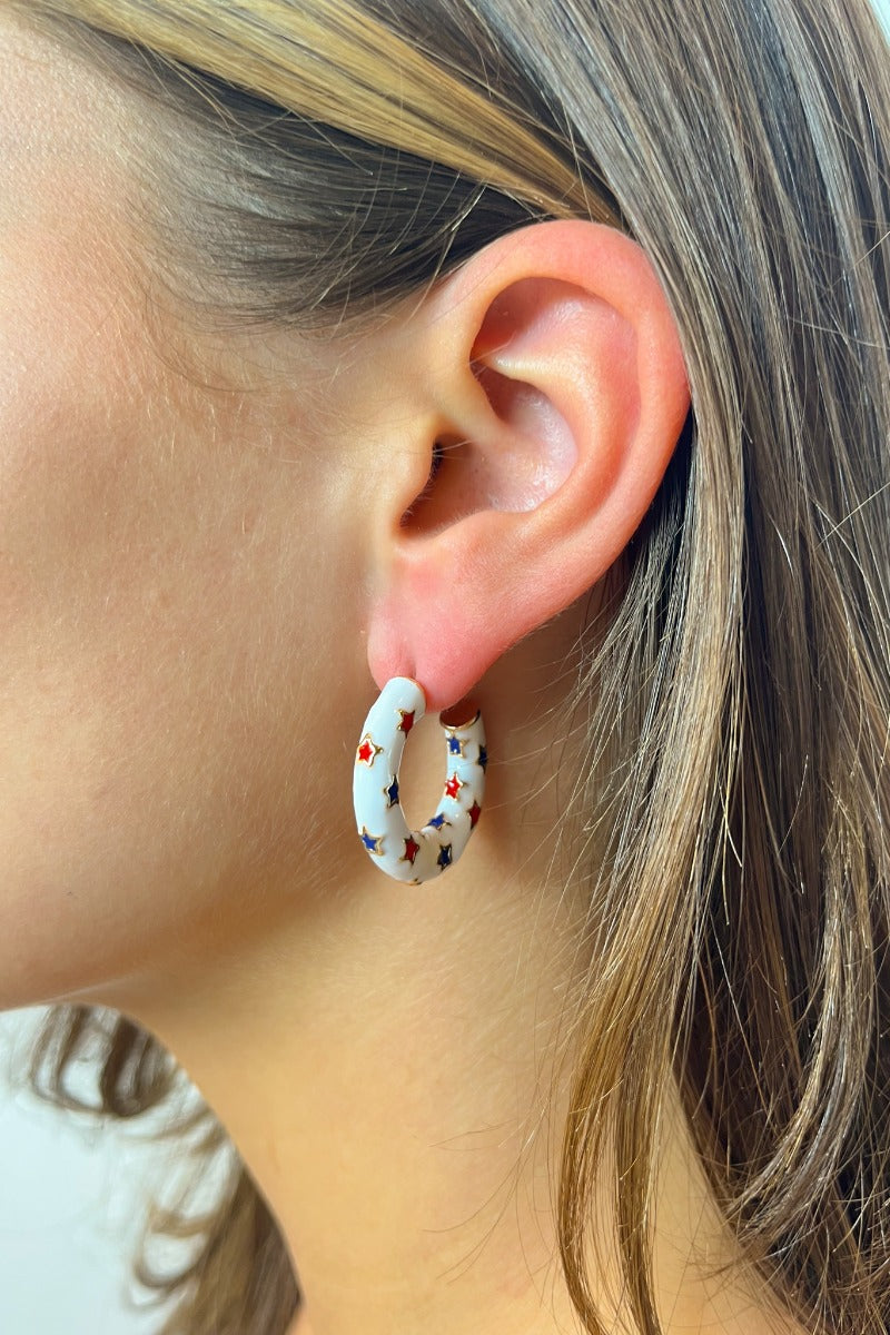 Side view of model wearing the Star Gazing Earrings in White which features small open white hoop with red and blue stars design outlined in gold details.