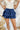 Frontal view of the Maeve Skort that features a blue satin material, a thick elastic waist band with a tie detail, a ruffle tiered design, a shorts lining, and a mini length.