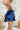 Side view of the Maeve Skort that features a blue satin material, a thick elastic waist band with a tie detail, a ruffle tiered design, a shorts lining, and a mini length.