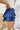 Side view of the Maeve Skort that features a blue satin material, a thick elastic waist band with a tie detail, a ruffle tiered design, a shorts lining, and a mini length.