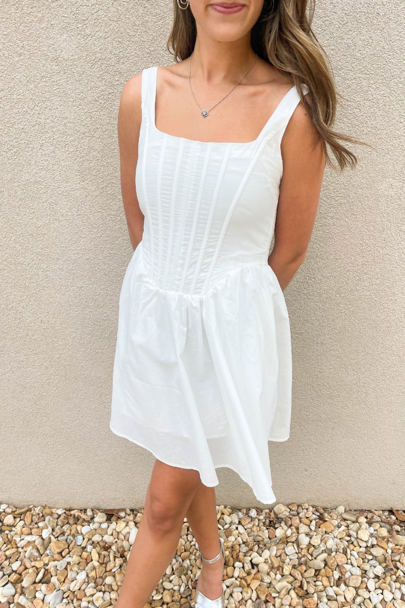 Close up view of model wearing the Prepare To Swoon Dress which features white fabric, mini length, white lining, pockets on each side, boning upper details, square neckline, thick straps, sleeveless and monochromatic back zipper with hook closure.
