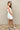 Full body side view of model wearing the Prepare To Swoon Dress which features white fabric, mini length, white lining, pockets on each side, boning upper details, square neckline, thick straps, sleeveless and monochromatic back zipper with hook closure.