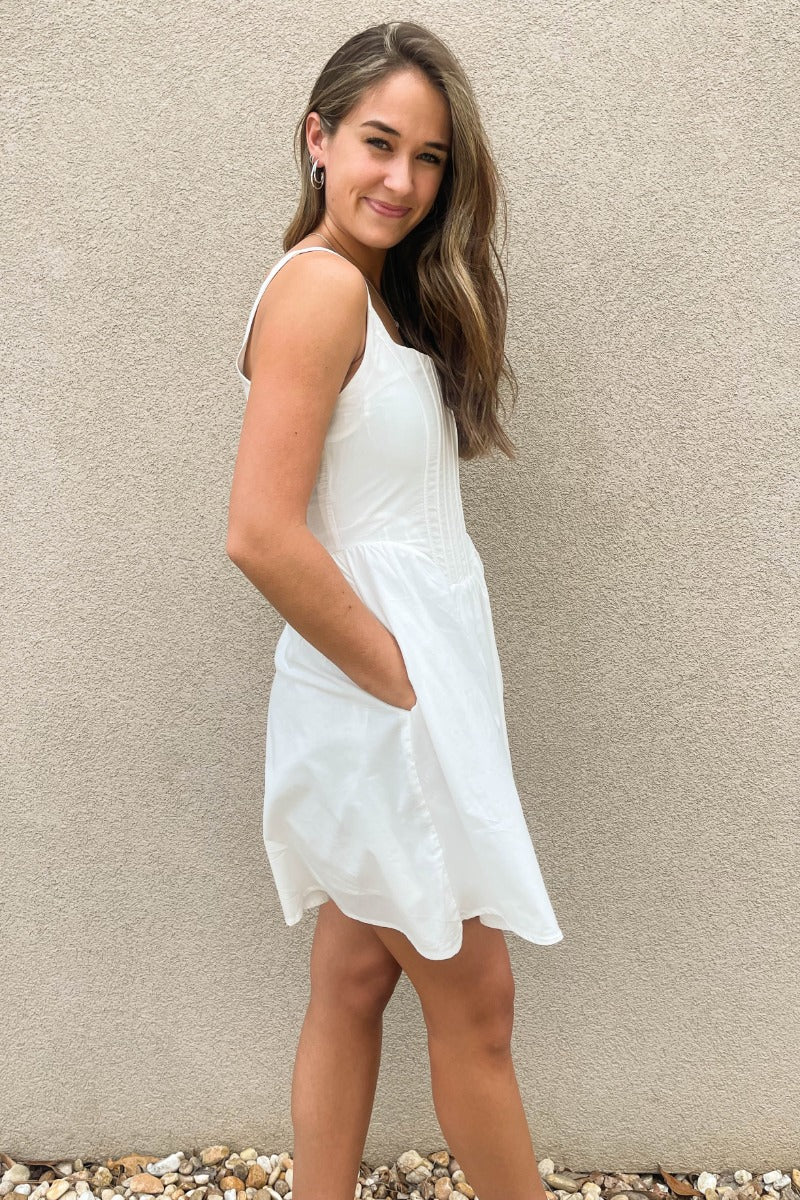 Side view of model wearing the Prepare To Swoon Dress which features white fabric, mini length, white lining, pockets on each side, boning upper details, square neckline, thick straps, sleeveless and monochromatic back zipper with hook closure.