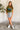 Full body front view of model wearing the Bronco Graphic Tee that has olive green knit fabric, short sleeves, a round neckline and a vintage bronco with a multi color background. Graphic says "Bronco.