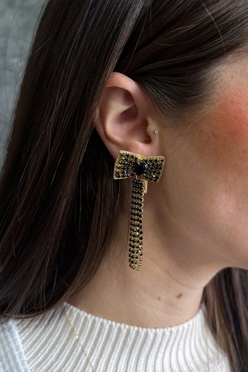 Side view of model wearing the Black Rhinestones Bow Shaped Dangle Earring which features bow shaped studs with black stones and black stone fringe details with gold trim.