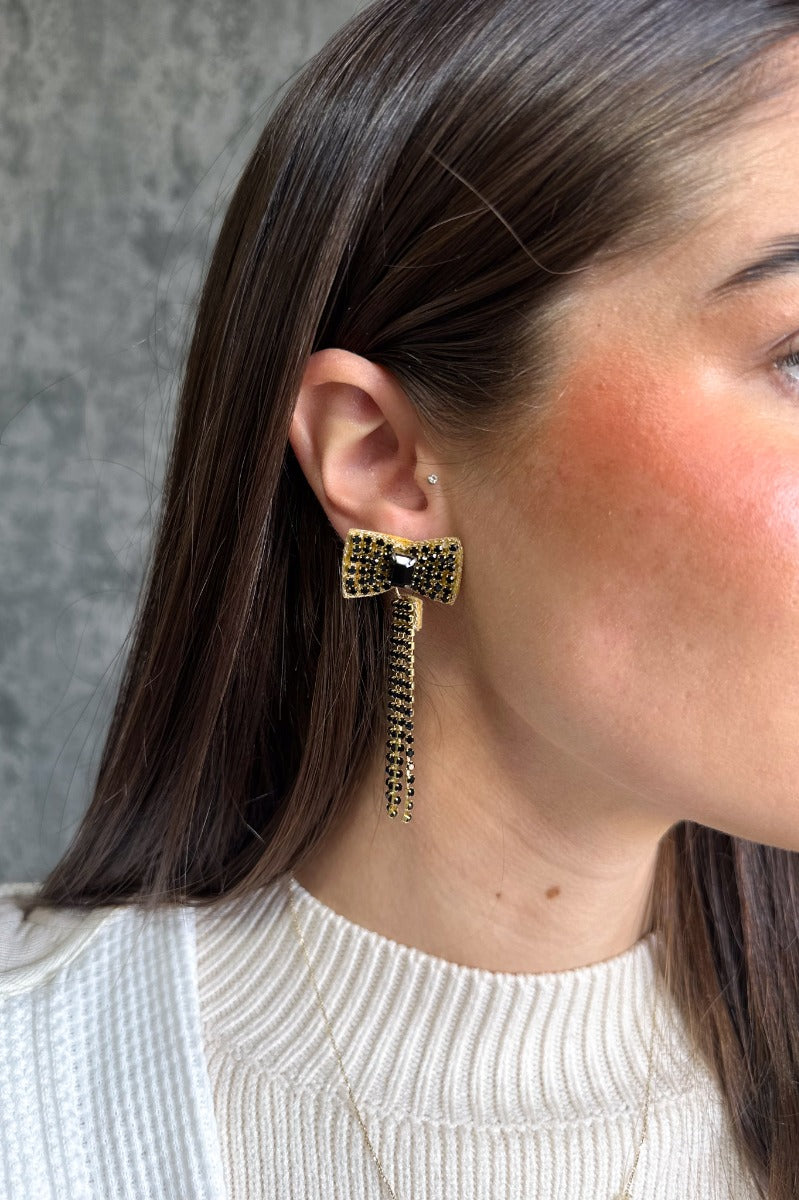 Side view of model wearing the Black Rhinestones Bow Shaped Dangle Earring which features bow shaped studs with black stones and black stone fringe details with gold trim.