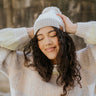 Front view of model wearing the Stay Cozy Beanie in Ivory, that has ivory sherpa fabric, taupe cable knit trim, a taupe pom pom, and a brown patch that says "C.C"