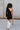 Full body back view of model wearing the Shelby Color Block Dress which features black and brown color blocked ribbed fabric, one long sleeve and one adjustable spaghetti strap.