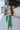Side view of model wearing the Feeling Lucky Midi Dress which features green fabric with a metallic sheen, a green lining, adjustable thin straps that tie across the back, and a side zipper.
