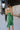 Back view of model wearing the Feeling Lucky Midi Dress which features green fabric with a metallic sheen, a green lining, adjustable thin straps that tie across the back, and a side zipper.