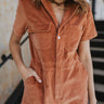 Close up view of the Talk To Me Jumpsuit which features rust corduroy fabric, a front zipper with a silver button closure, 2 chest pockets, 2 side pockets, a collared neckline and short sleeves.