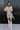 Full body front view of model wearing the Pop The Champagne Dress, that has pleated champagne-colored fabric with metallic gold stripes, long sleeves with buttoned cuffs, a button-up front with a collared neckline, and a matching detachable belt