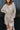 Close front view of model wearing the Pop The Champagne Dress, that has pleated champagne-colored fabric with metallic gold stripes, long sleeves with buttoned cuffs, a button-up front with a collared neckline, and a matching detachable belt