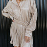 Close front view of model wearing the Pop The Champagne Dress, that has pleated champagne-colored fabric with metallic gold stripes, long sleeves with buttoned cuffs, a button-up front with a collared neckline, and a matching detachable belt