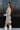 Side view of model wearing the Pop The Champagne Dress, that has pleated champagne-colored fabric with metallic gold stripes, long sleeves with buttoned cuffs, a button-up front with a collared neckline, and a matching detachable belt
