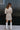 Full body back view of model wearing the Pop The Champagne Dress, that has pleated champagne-colored fabric with metallic gold stripes, long sleeves with buttoned cuffs, a button-up front with a collared neckline, and a matching detachable belt