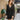 Front view of model wearing the Promise Me Dress, that has lined black fabric with a surplice neckline, a smocked waist, a tiered skirt, and semi-sheer long sleeves with a slight sheen and elastic wrists