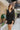 Front view of model wearing the Promise Me Dress, that has lined black fabric with a surplice neckline, a smocked waist, a tiered skirt, and semi-sheer long sleeves with a slight sheen and elastic wrists