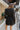 Back view of model wearing the Promise Me Dress, that has lined black fabric with a surplice neckline, a smocked waist, a tiered skirt, and semi-sheer long sleeves with a slight sheen and elastic wrists