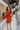 Full body side view of model wearing the Falling Fast Dress, that has rust-colored fabric, a v-neckline, ruffle details on the shoulders, long balloon sleeves with elastic wrists, and side pockets