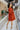Full body back view of model wearing the Falling Fast Dress, that has rust-colored fabric, a v-neckline, ruffle details on the shoulders, long balloon sleeves with elastic wrists, and side pockets