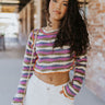 Front view of model wearing the So Over It Sweater, that has a beige, purple, blue, and olive digital striped pattern with orange lettuce trim, a cropped waist, long sleeves, and a round neckline.