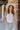 Front view of model wearing the Talia Bodysuit In White, that has white ribbed fabric, a round neckline, and a thong bottom with hook closures. Worn with jeans.