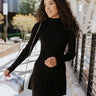 Front view of model wearing the Elodie Dress in Black, which features black ribbed fabric, a high neck with lettuce trim, long sleeves, and a flared skirt