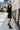 Full body side view of model wearing the Elodie Dress in Black, which features black ribbed fabric, a high neck with lettuce trim, long sleeves, and a flared skirt