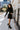 Full body side view of model wearing the Elodie Dress in Black, which features black ribbed fabric, a high neck with lettuce trim, long sleeves, and a flared skirt