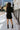 Full body back view of model wearing the Elodie Dress in Black, which features black ribbed fabric, a high neck with lettuce trim, long sleeves, and a flared skirt