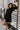 Front view of model wearing the Elodie Dress in Black, which features black ribbed fabric, a high neck with lettuce trim, long sleeves, and a flared skirt