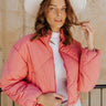 Front view of model wearing the Aspen Puffer Jacket in Pink, that has pink puffer fabric with diamond stitching. a high neck, a front zipper, two front pockets, a cropped waist, and long sleeves. Worn unzipper over white top.