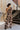 Full body back view of model wearing the Good Vibes Floral Top, that has brown light weight fabric with taupe flowers pattern, one shoulder with a long sleeve, a cropped waist, and a brown zipper on the left side. Tucked into matching pants.
