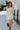 Full body front view of model wearing the Elodie Dress in Cream, which features cream ribbed fabric, a high neck with lettuce trim, long sleeves, and a flared skirt