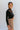 Side view of model wearing the Last Call Bodysuit, which features black velvet with a mesh neckline, long sleeves, and a cheeky bottom. Worn with plaid skirt.