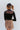 Back view of model wearing the Last Call Bodysuit, which features black velvet with a mesh neckline, long sleeves, and a cheeky bottom. Worn with plaid skirt.