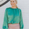 Front view of model wearing the Lost In Your Eyes Top, which features a green satin fabric, long sleeves, a round high neckline with pleating, and an elastic waist.
