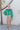 Front view of model wearing the Lost In Your Eyes Top, which features a green satin fabric, long sleeves, a round high neckline with pleating, and an elastic waist.