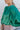 Close-up side view of model wearing the Lost In Your Eyes Top, which features a green satin fabric, long sleeves, a round high neckline with pleating, and an elastic waist.