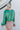 Back view of model wearing the Lost In Your Eyes Top, which features a green satin fabric, long sleeves, a round high neckline with pleating, and an elastic waist.