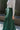 Close-up side view of model wearing the Nothing But Love Pants, that have dark green fabric with inverted front pleating, an attached tie belt, wide flowy legs, and a side zipper. Worn with white top.